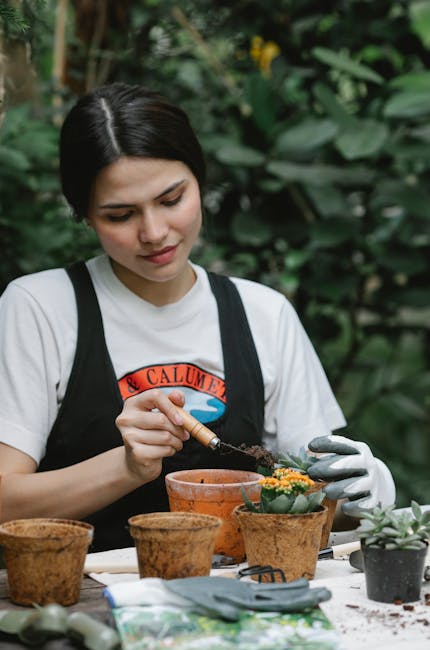 A woman with short dark hair, wearing a white t-shirt and black apron, is engaged in garden planting work outdoors. She is using a wooden-handled tool to carefully transplant or pot small flowering plants into terracotta pots on a white work surface. The pots vary in size and are arranged in front of her, with some containing soil and young plants, while others are empty or partially filled. In the background, lush green foliage, including large-leaved plants and possibly small shrubs or trees, provides a dense natural setting. The scene is well-lit with natural outdoor light, emphasizing the textures of the moist soil, the smooth terracotta, and the vibrant green leaves. Gardeners Bermondsey’s expertise is evident in the careful attention to detail, suggesting activities like container gardening, plant care, or preparing plants for a small yard or garden border as part of their landscaping work.