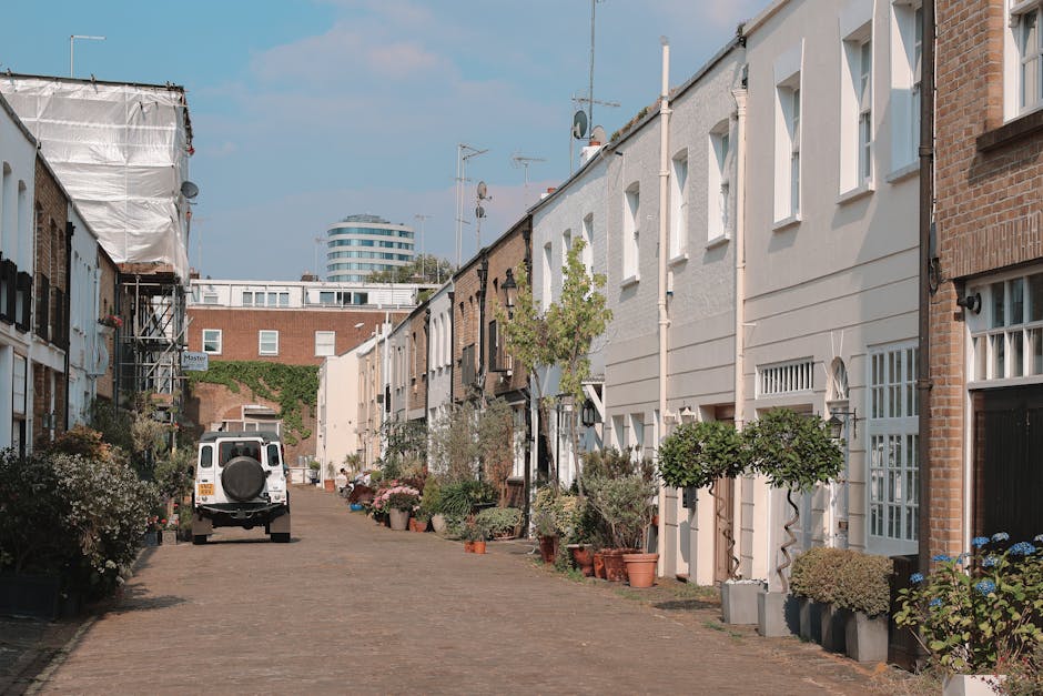 A quiet urban garden scene in Bermondsey featuring a narrow cobblestone pathway bordered by a mix of small trees, potted plants, and neatly trimmed shrubs. On one side, there are modern white and brick terraced houses with multiple windows and front doors, some decorated with climbing plants and planters. The pathway is lined with various garden pots containing flowering plants and small trees, suggesting ongoing gardening or landscape maintenance. A white utility vehicle with a spare tire mounted on the back is parked along the street, indicating recent or upcoming garden servicing by Gardeners Bermondsey. In the background, there are taller buildings and a partially clear blue sky, creating a blend of residential and urban landscape. The scene emphasizes tidy, well-maintained outdoor spaces and the practical aspects of garden and landscape care in an inner-city setting, illustrating routine garden maintenance and planting activities aligned with the topic of access issues for Bermondsey garden services.